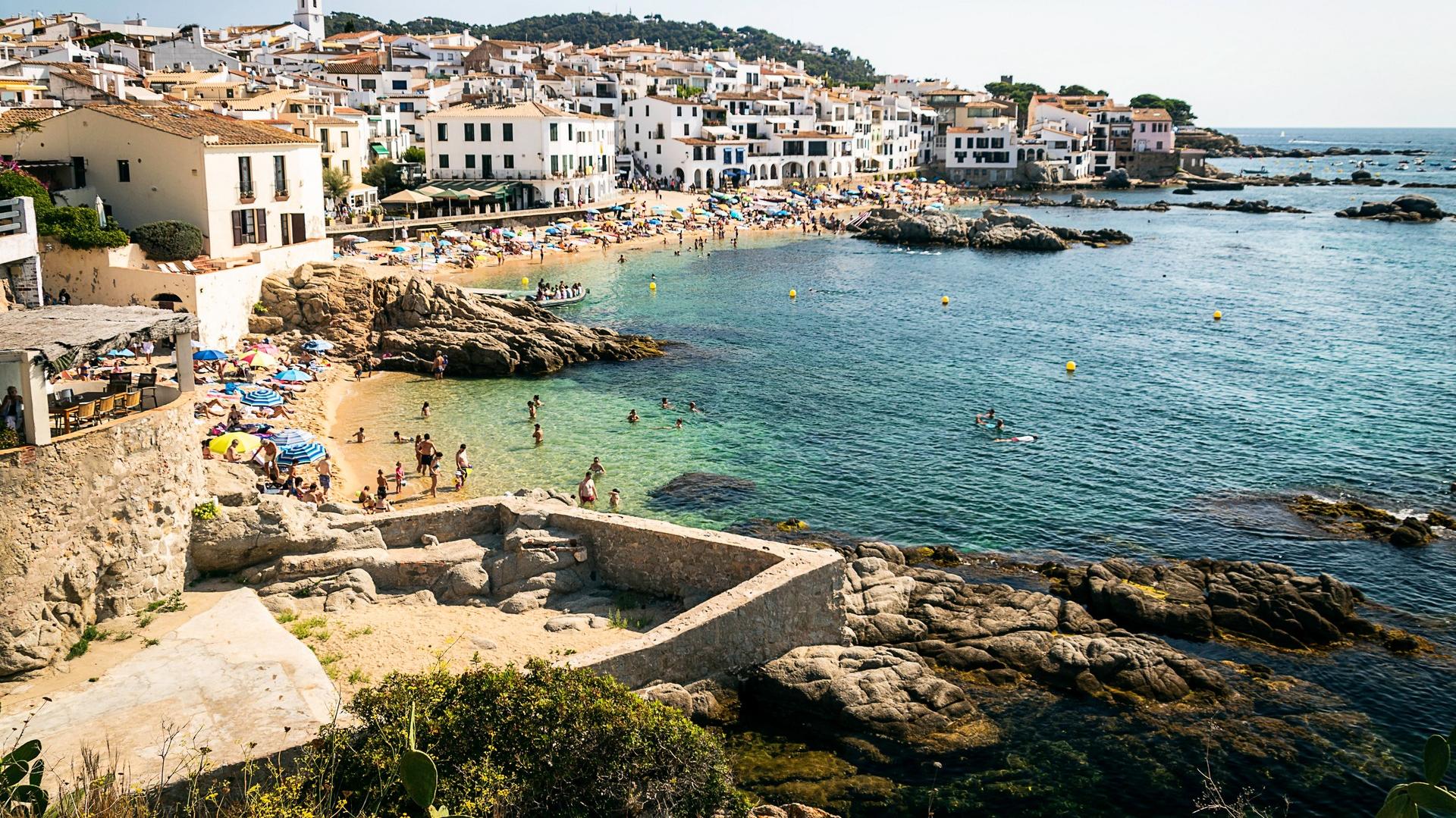 Beach with a lot of people and architecture in Calella de Palafrugell on a day with cloudy weather